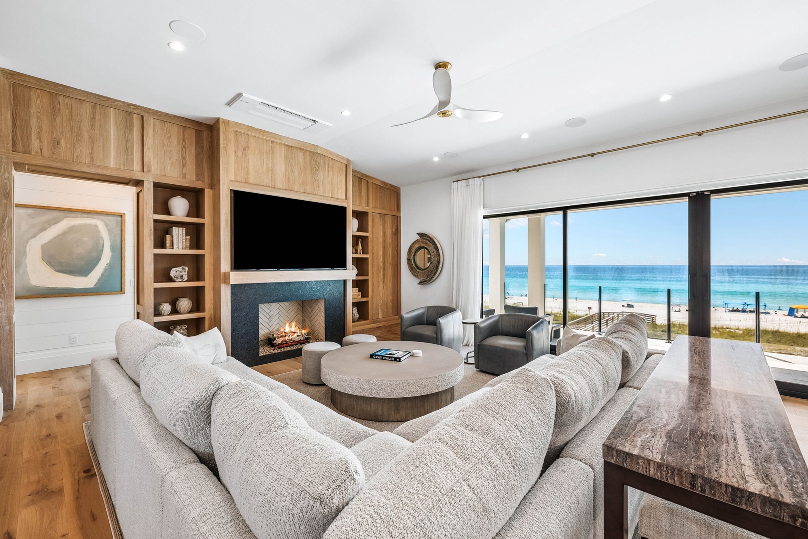 Sink into plush seating and gaze out at stunning ocean views framed by floor-to-ceiling windows, where warm wood tones and a cozy fireplace create the perfect beachside retreat.