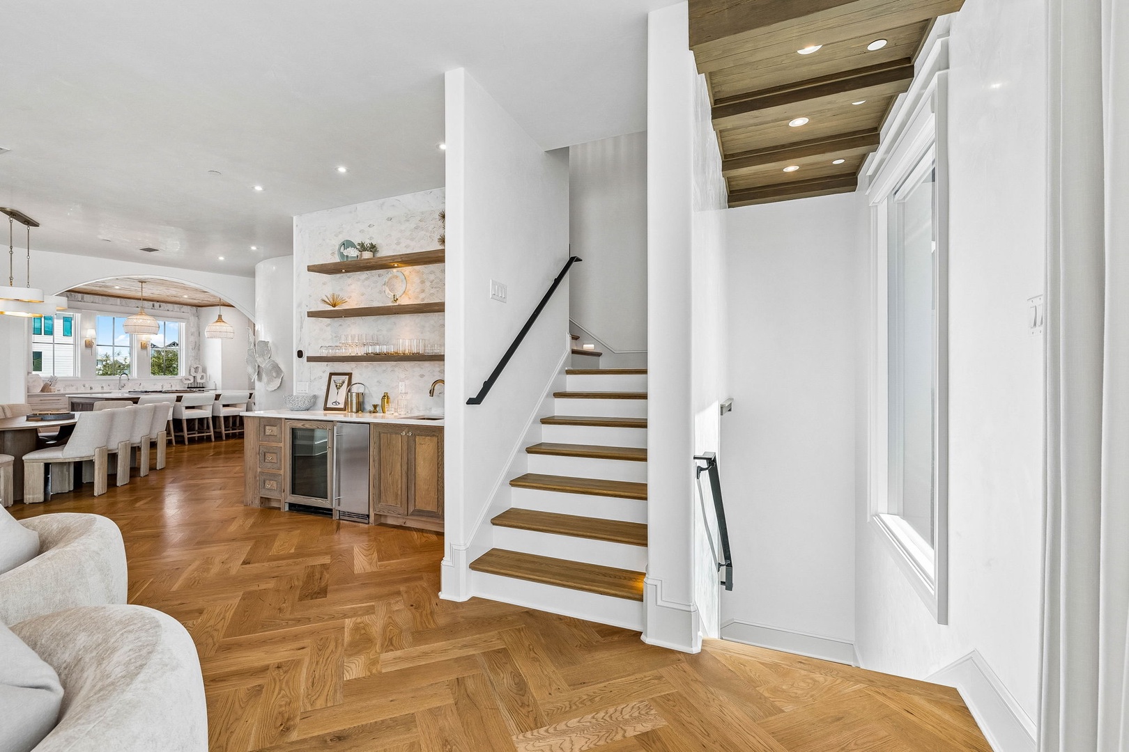 Bright, open-plan living flows seamlessly from your cozy seating area through a sun-drenched dining space to the kitchen, all connected by warm herringbone floors and architectural wood ceiling accents.