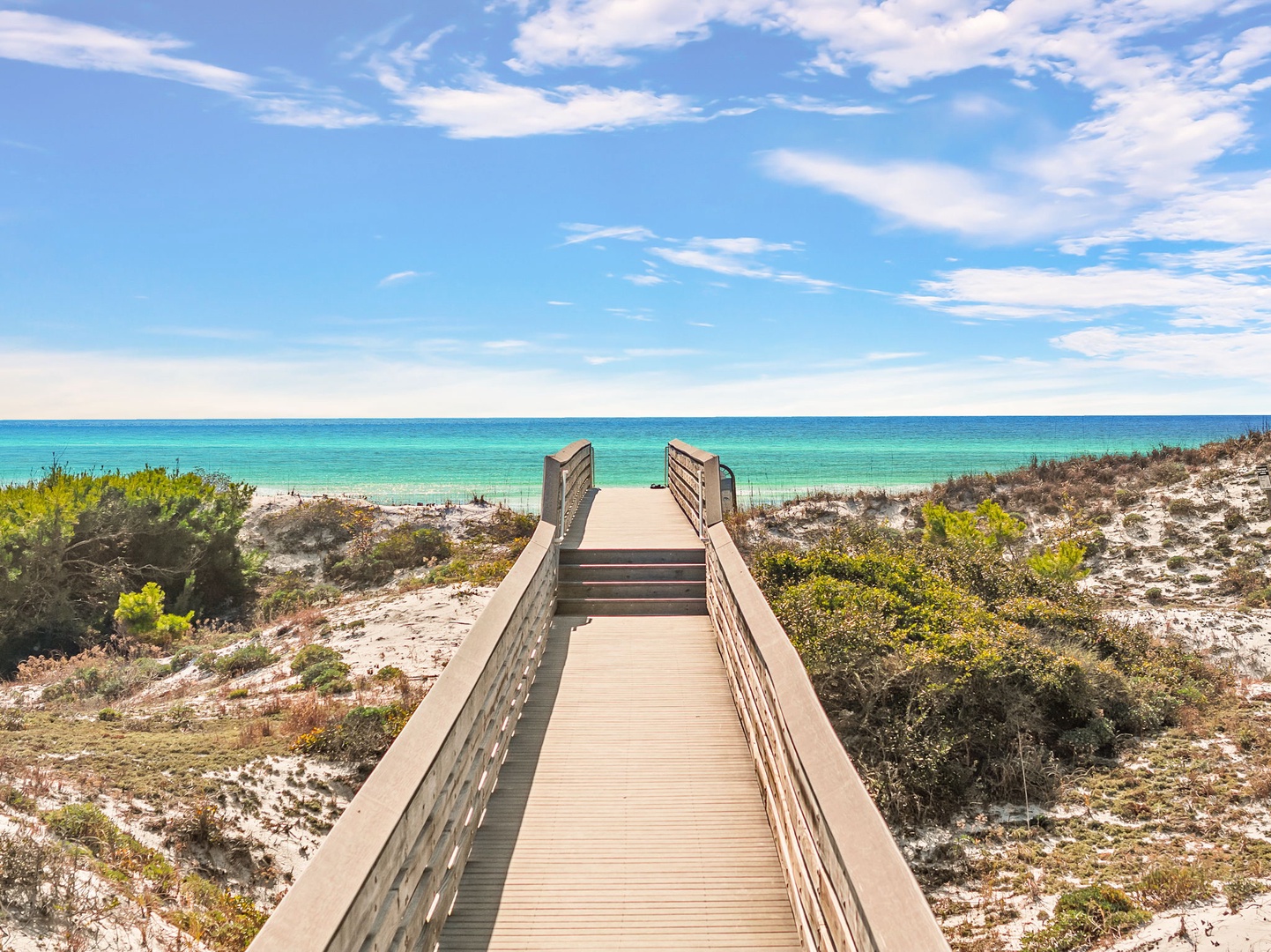 Wooden boardwalk leads across sandy dunes to pristine turquoise waters and white sand beach under brilliant blue skies.