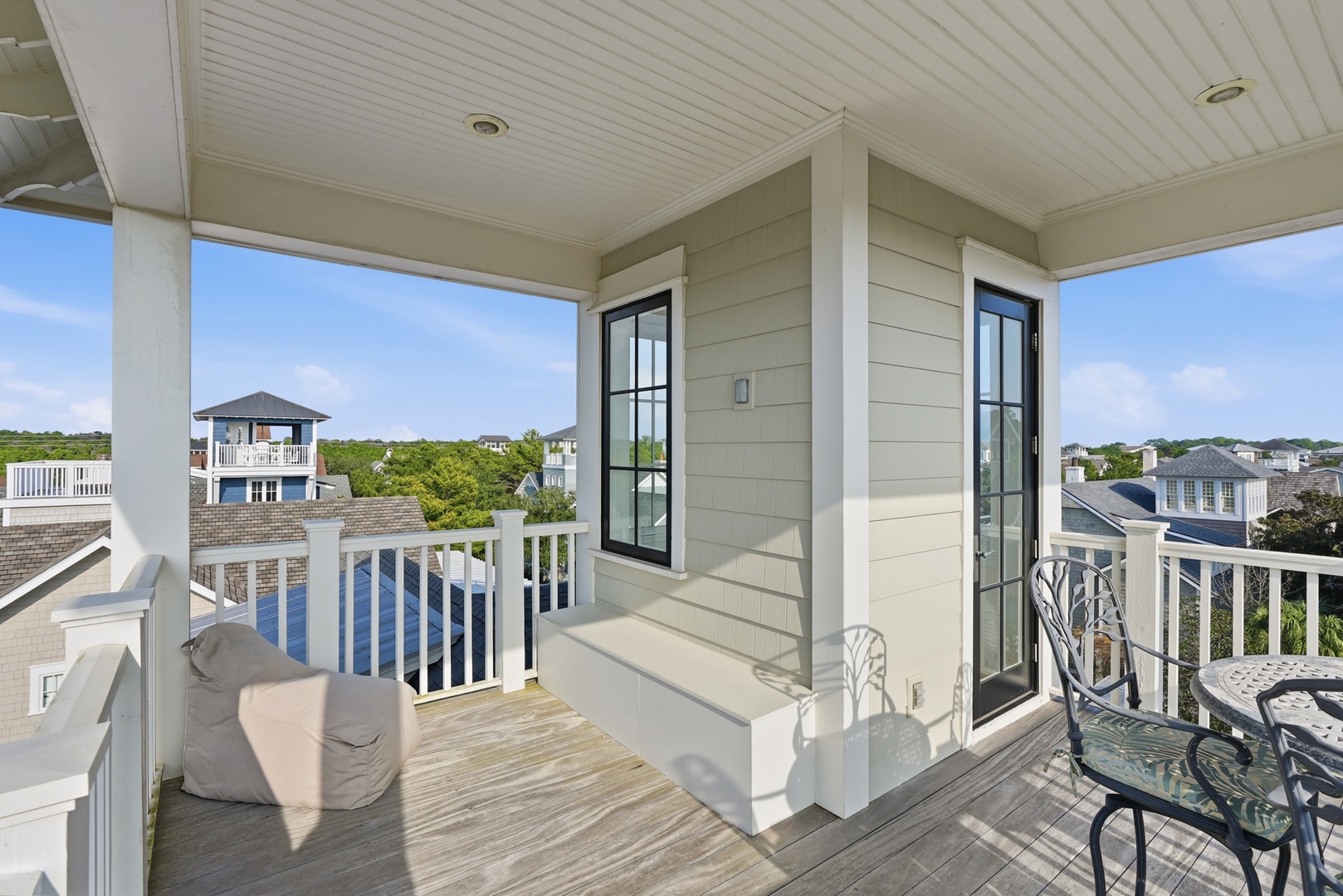Elevated balcony overlooking a coastal neighborhood, framed by white railings and black-trimmed French doors. Rooftop views extend across tree-lined residential streets and neighboring homes.