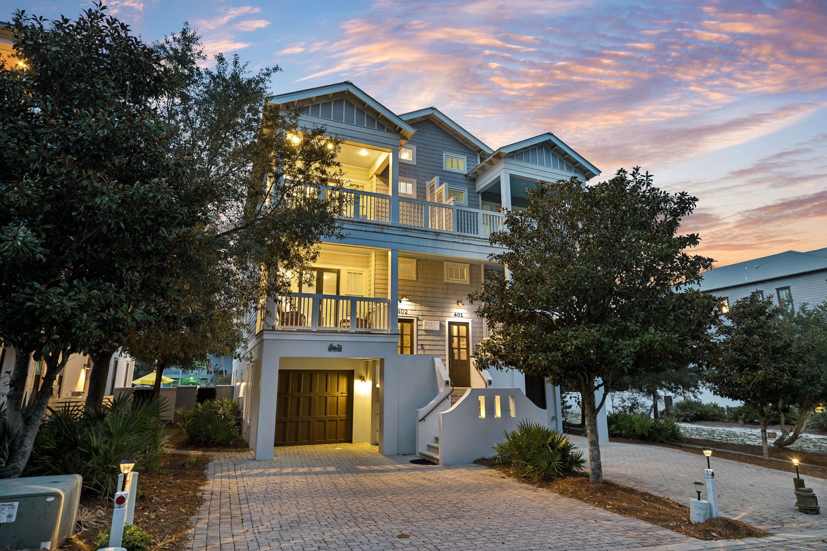 A coastal-style home framed by native trees and greenery, featuring multiple balconies and warm evening lighting, located in a peaceful neighborhood setting.