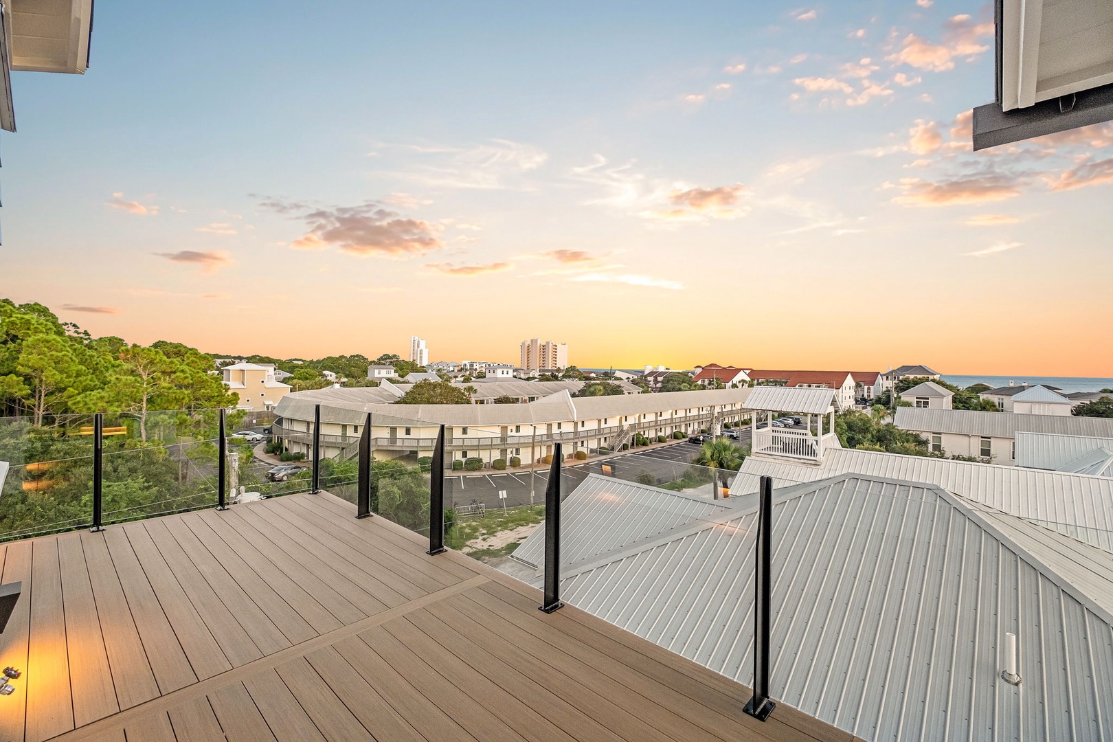 Elevated balcony overlooks the neighborhood rooftops and distant coastline, with a sunset glow painting the sky in soft coral and blue tones.