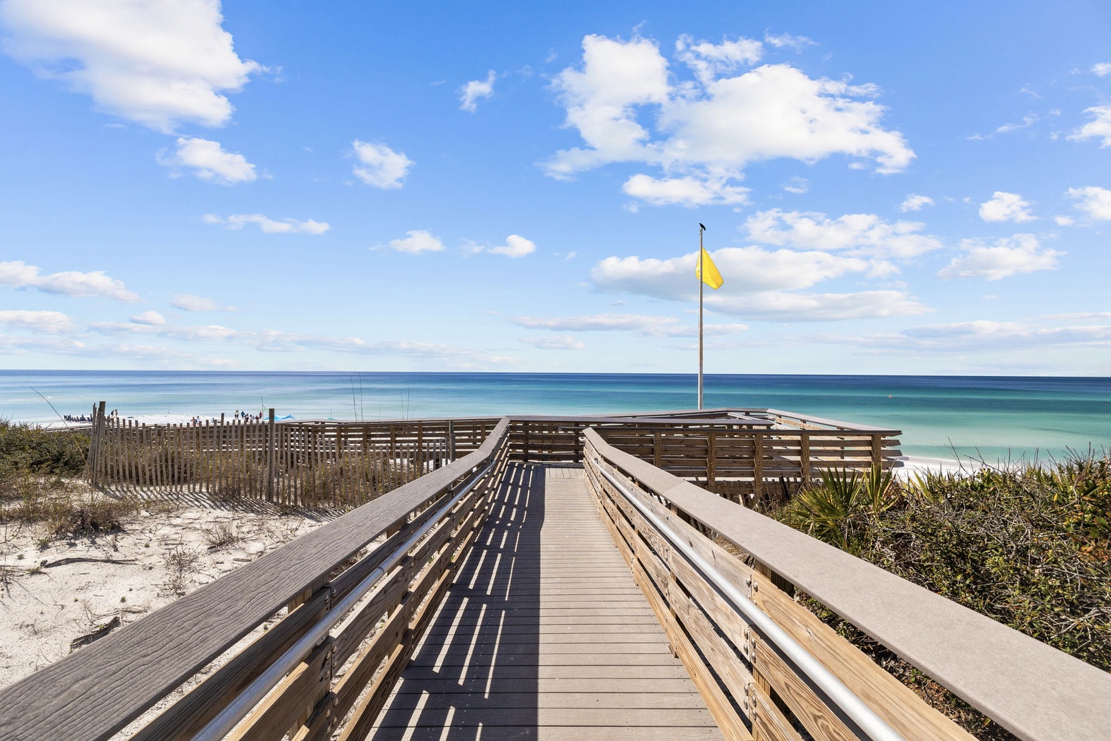 Wooden boardwalk leads across pristine dunes to turquoise waters and white sand beach under brilliant blue skies.