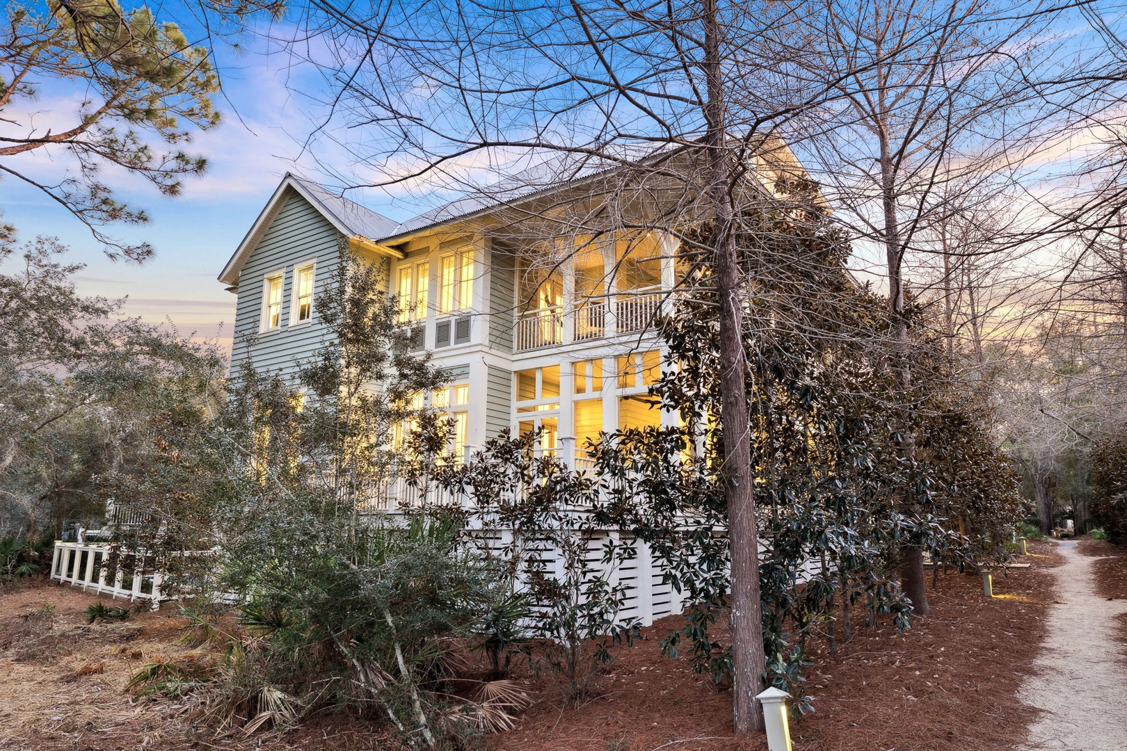 Charming two-story coastal home nestled among native trees with warm evening glow from windows.
