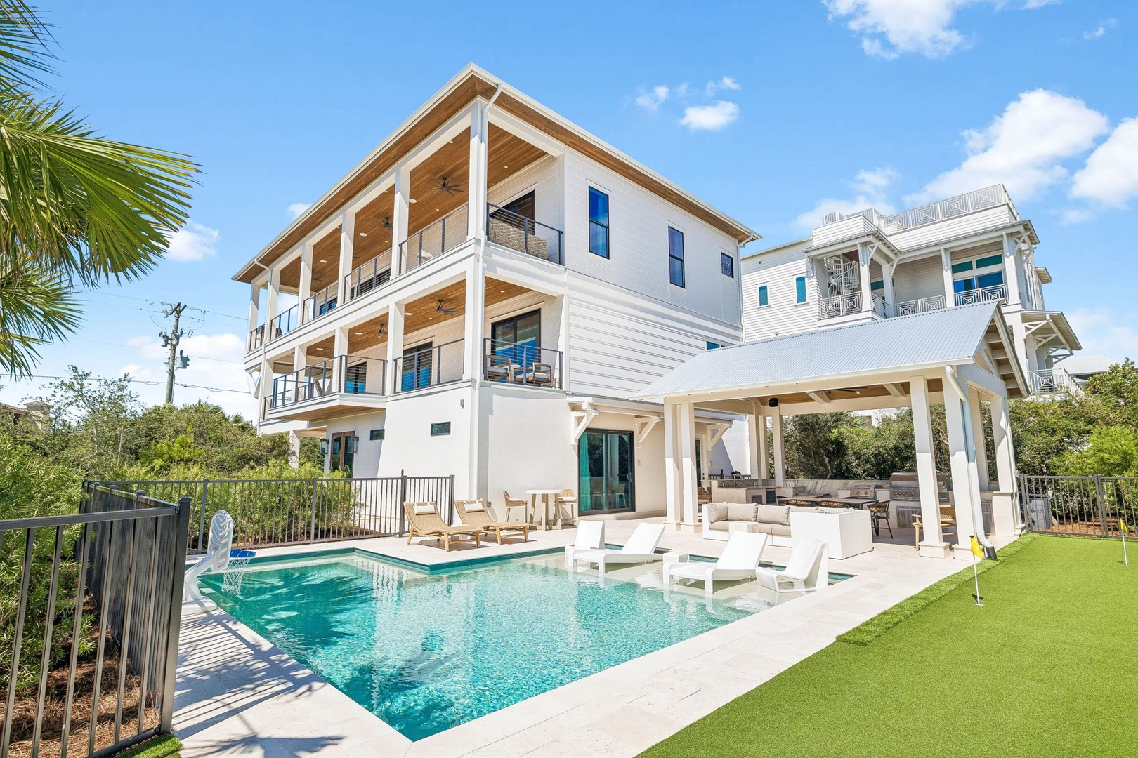 A contemporary three-story beachside property featuring a private pool, spacious patio with lounge seating, and manicured lawn surrounded by coastal palms and neighboring residences.