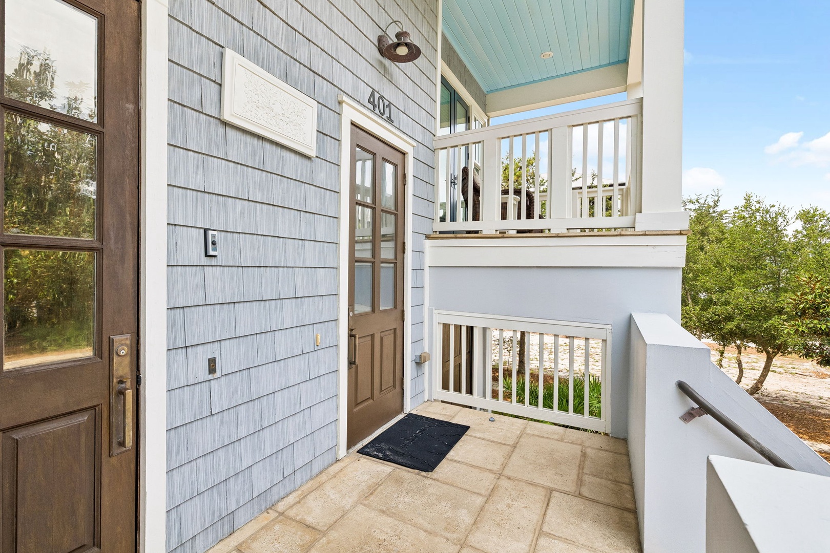 The front entrance features pale blue siding and a welcoming two-story porch, marking your arrival at this coastal-inspired retreat surrounded by greenery.