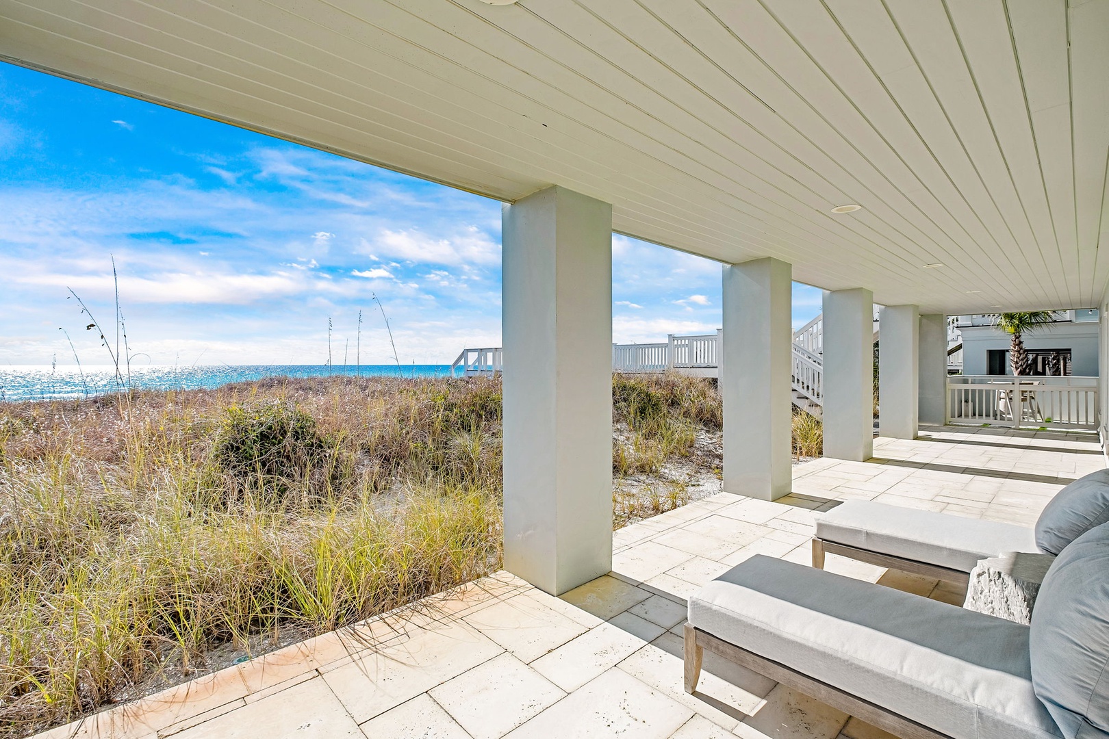 Covered beachfront terrace with lounge seating overlooking coastal dunes and sparkling ocean waters, steps from the sandy shore.