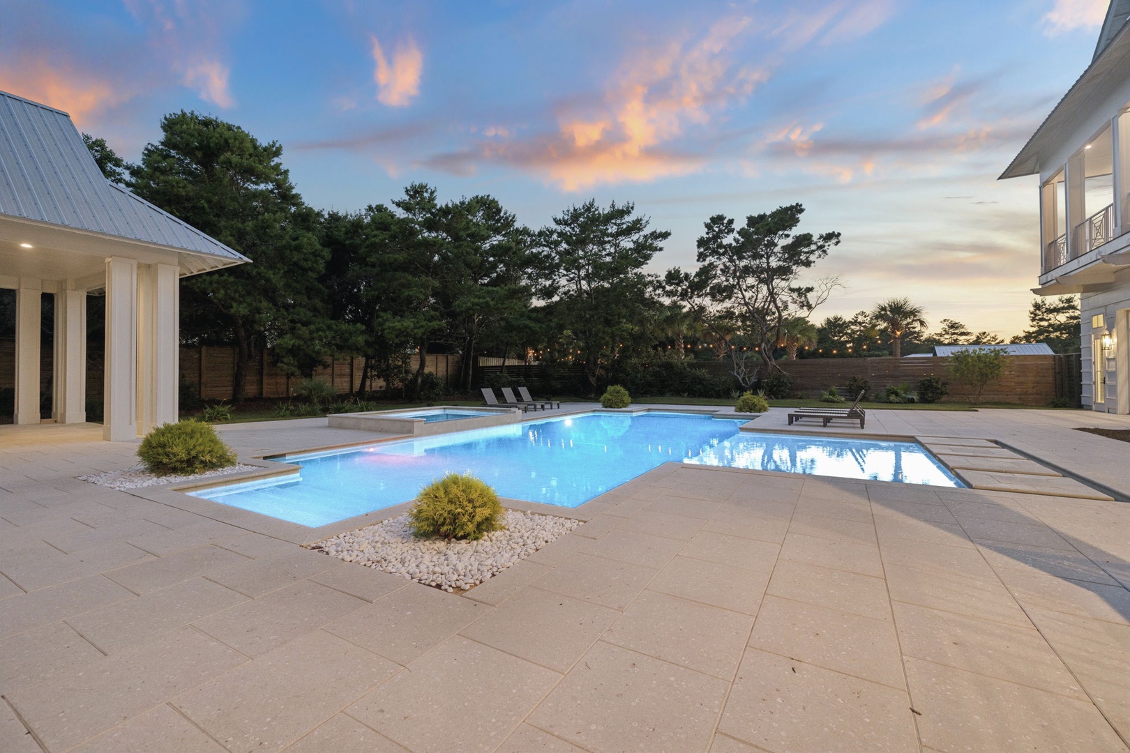 A stunning pool deck framed by mature trees and soft evening light. The spacious outdoor area features modern landscaping with decorative shrubs and stone accents, offering a peaceful retreat for relaxation.