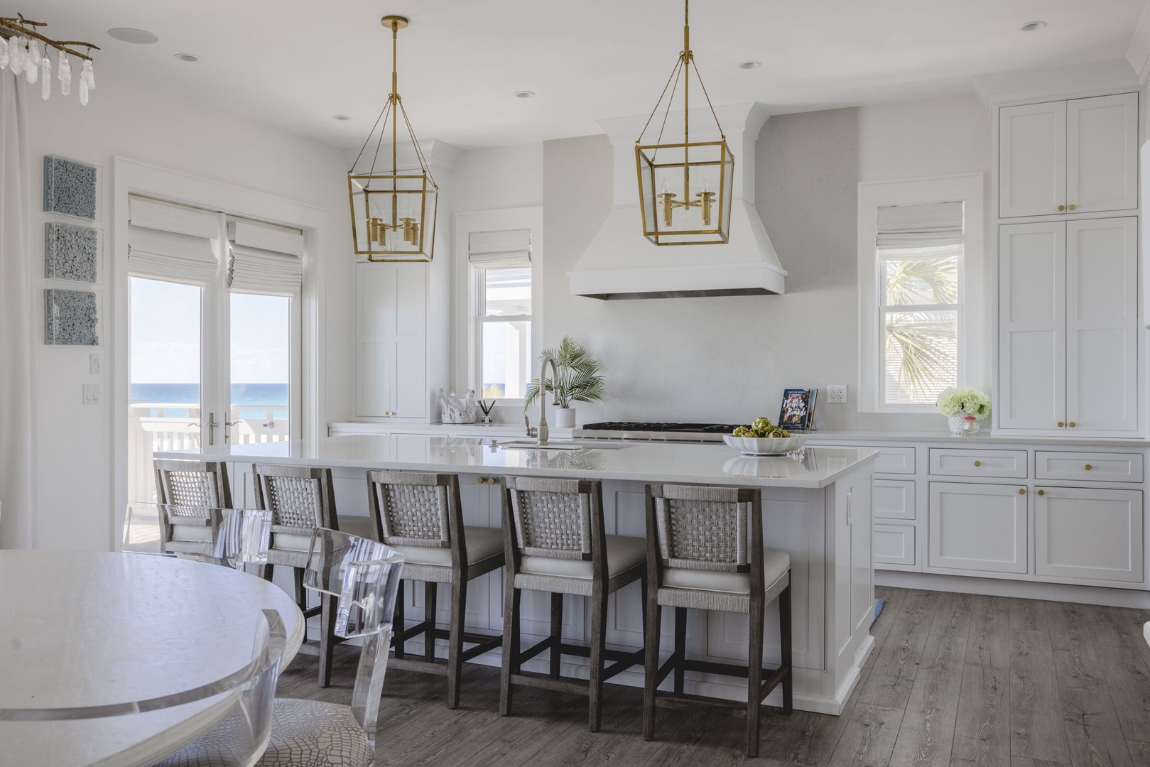 Your elegant coastal kitchen features marble countertops, a large island with bar seating, and stunning ocean views through French doors. Golden pendant lights add warmth to the bright space.