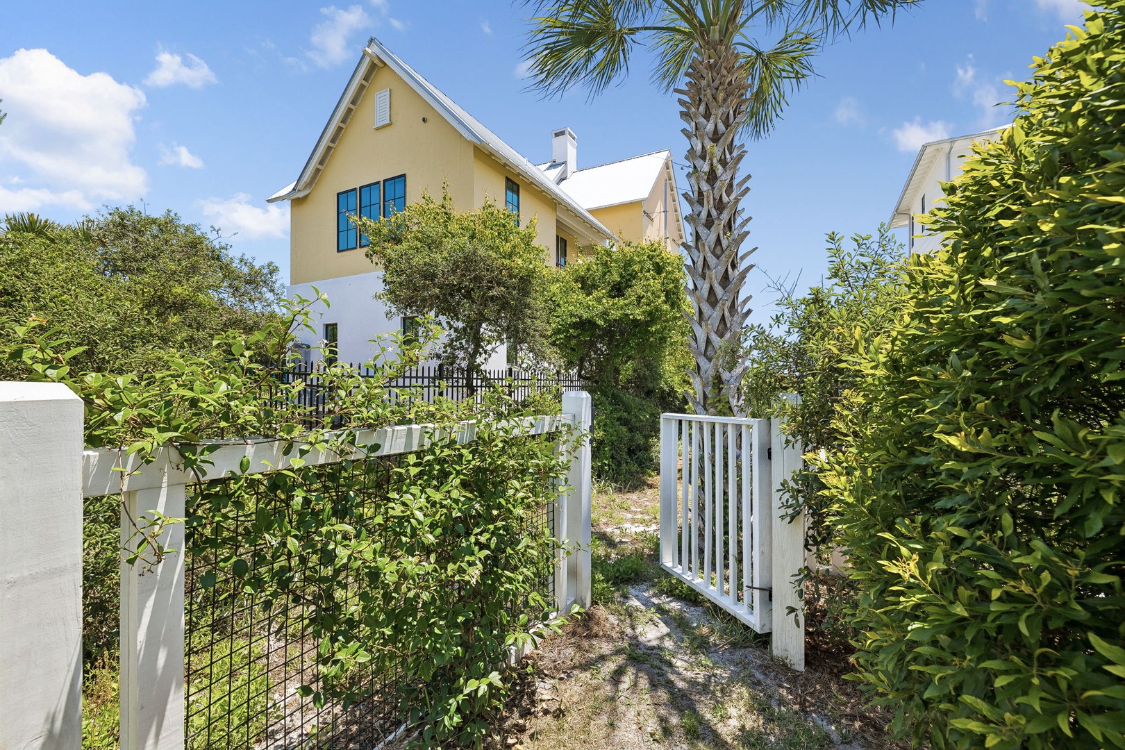A charming coastal-style vacation home features sunny yellow siding, surrounded by lush tropical palms and verdant greenery. The welcoming entrance gate opens to a private pathway.