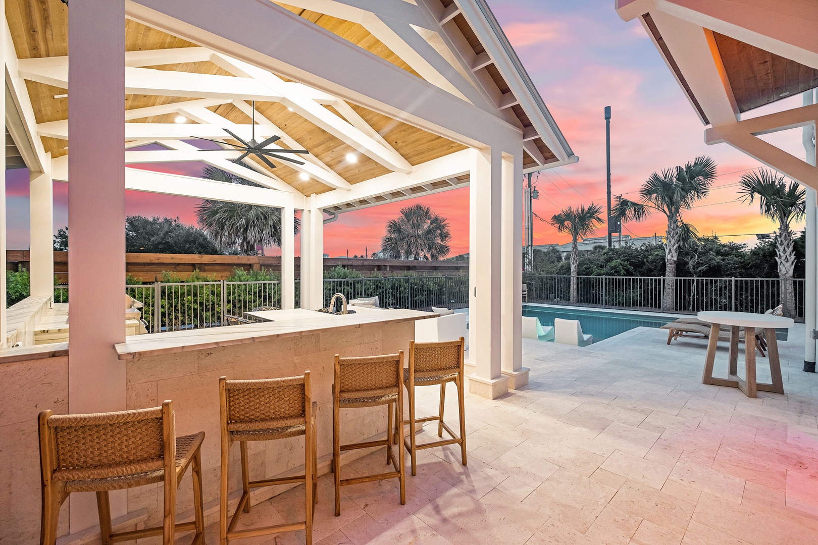 A covered outdoor bar area framing a stunning sunset over palm trees and a sparkling pool—an ideal setting for your evening gatherings.