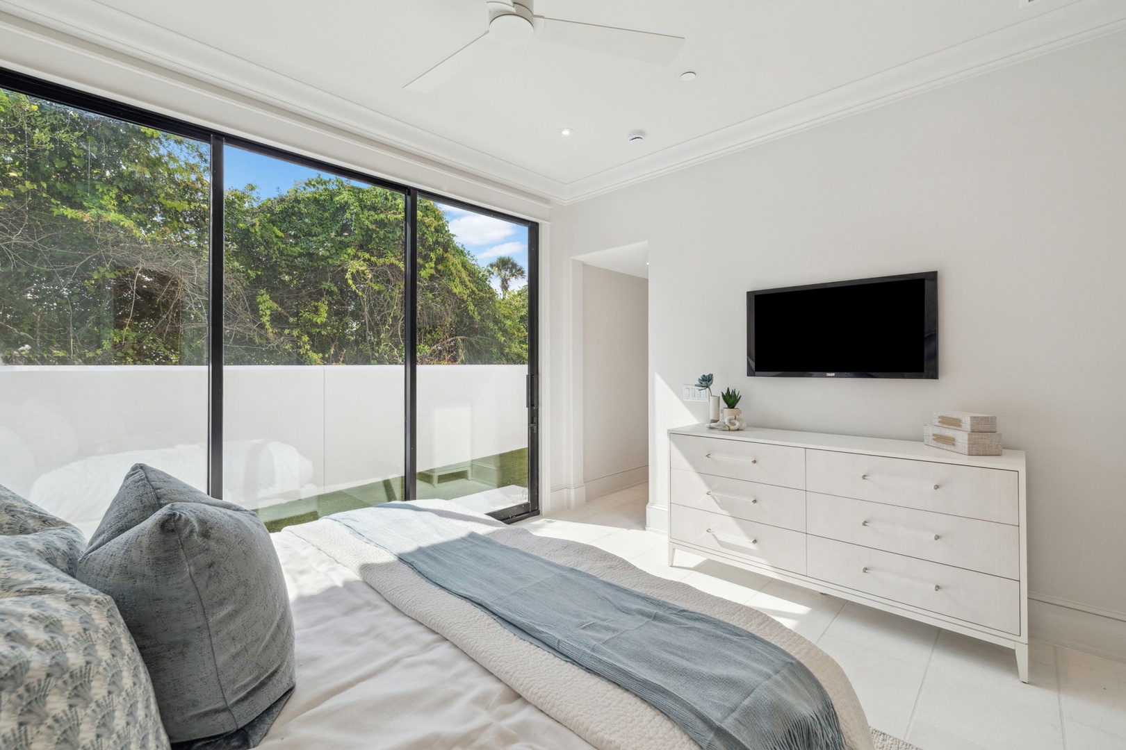 Wake to sunlight streaming through floor-to-ceiling windows overlooking lush greenery. Your serene bedroom features a plush bed, sleek storage, wall-mounted TV, and direct access to your private outdoor terrace.