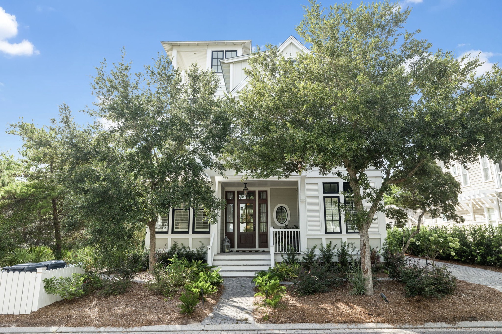 A charming white cottage with a welcoming front porch, framed by mature trees and lush greenery, offers a serene arrival to your coastal retreat.