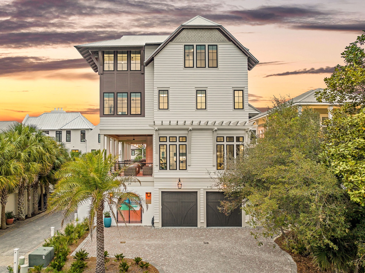 A stunning coastal home showcases modern architecture with palm trees and tropical landscaping under a dramatic sunset sky.
