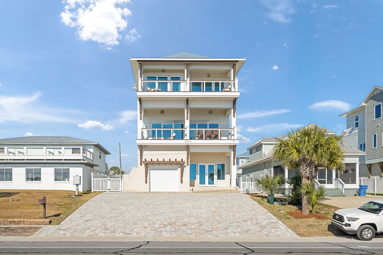 Modern three-story beach house with multiple balconies and spacious paver driveway in a coastal neighborhood.
