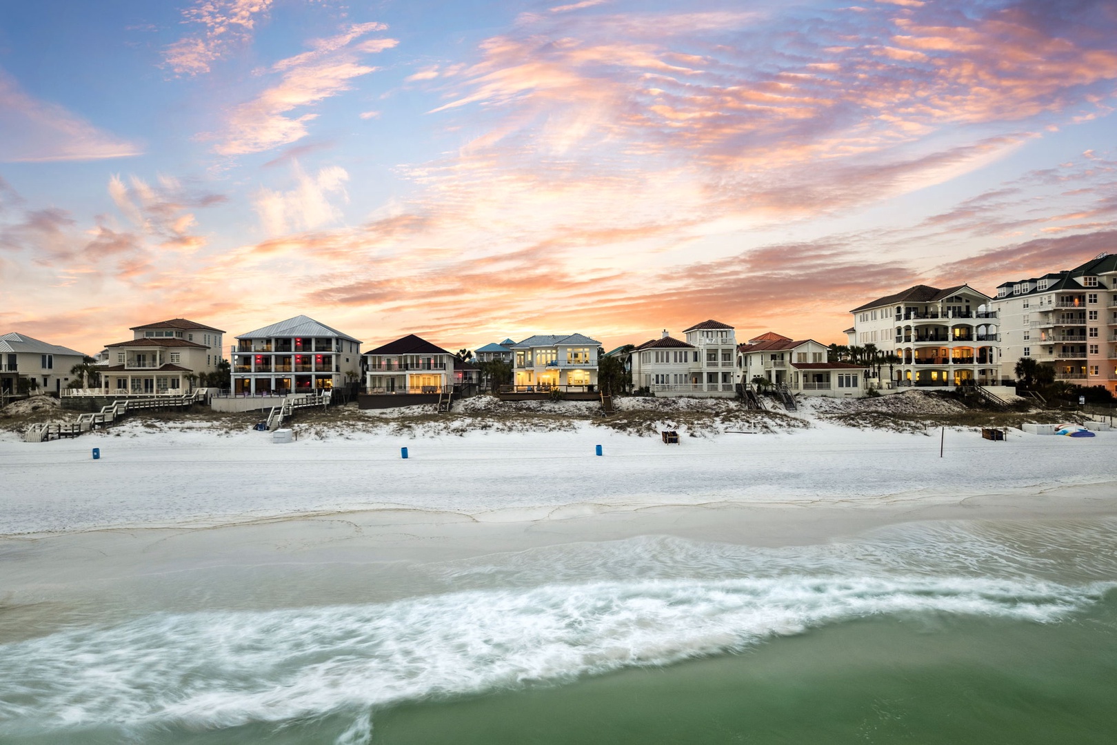 Beautiful beachfront homes line pristine white sand shores beneath a stunning sunset sky painted in soft pastels.