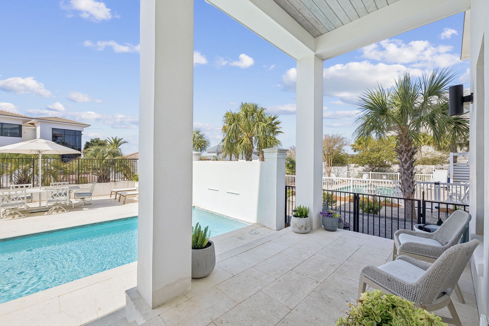 Your covered terrace features comfortable seating with stunning pool views, surrounded by tropical palms under sunny skies.