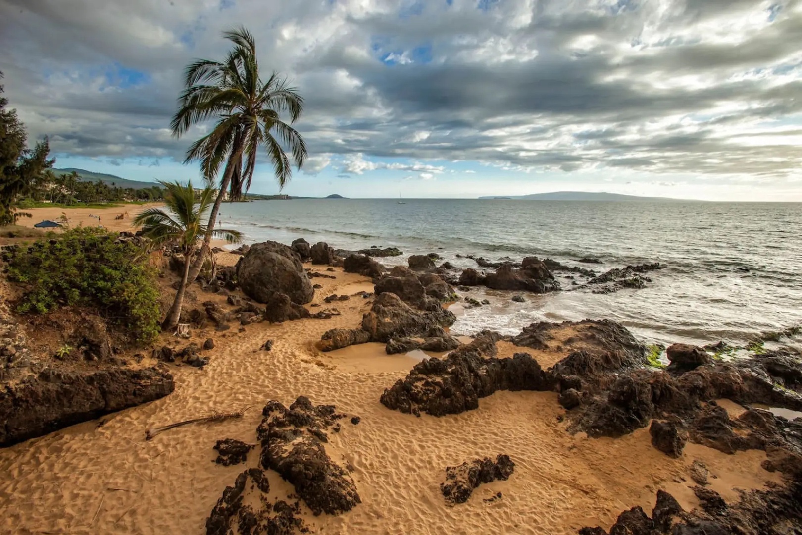 Kihei Vacation Rentals, Kalama Terrace P302 - Lava rock interrupts the sand where the beach curves, clouds rolling in behind the palms.