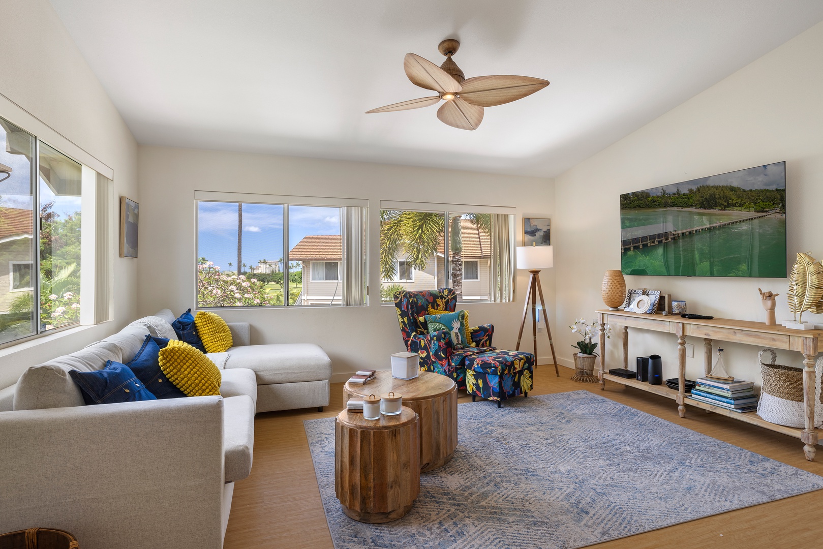 Relax in your bright, modern living room with tropical ceiling fan, comfortable seating, and glimpses of nearby homes through the windows.