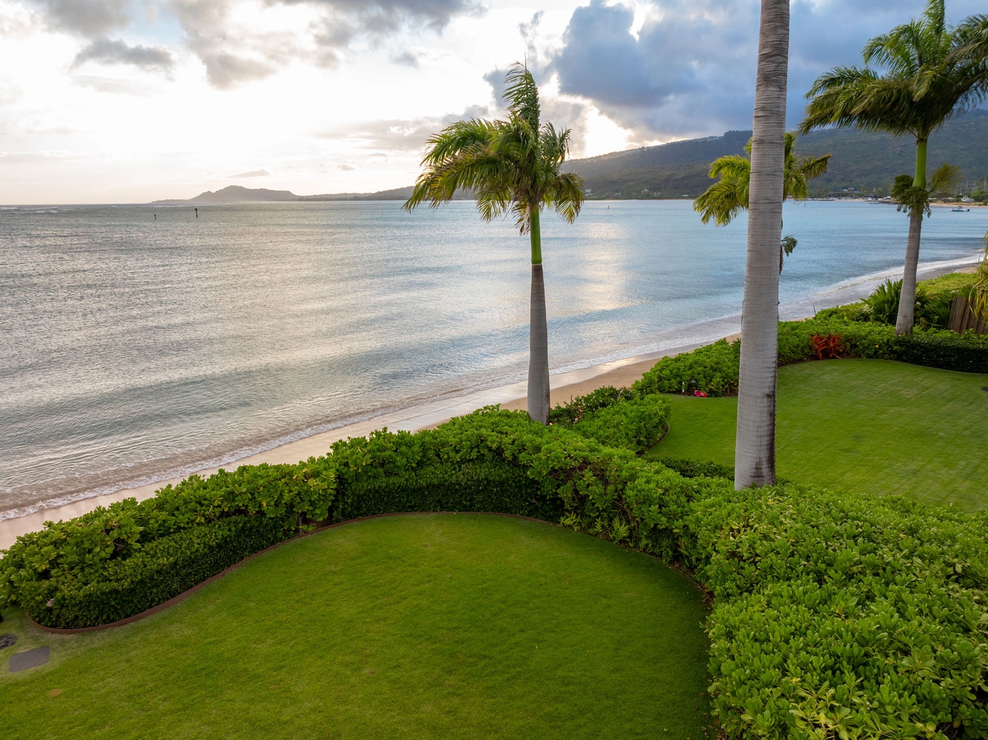 Honolulu Vacation Rentals, Maunalua Estate - Beachfront property featuring manicured lawns bordered by tropical palms, with pristine sands and calm turquoise waters stretching toward distant mountains under dramatic coastal skies.