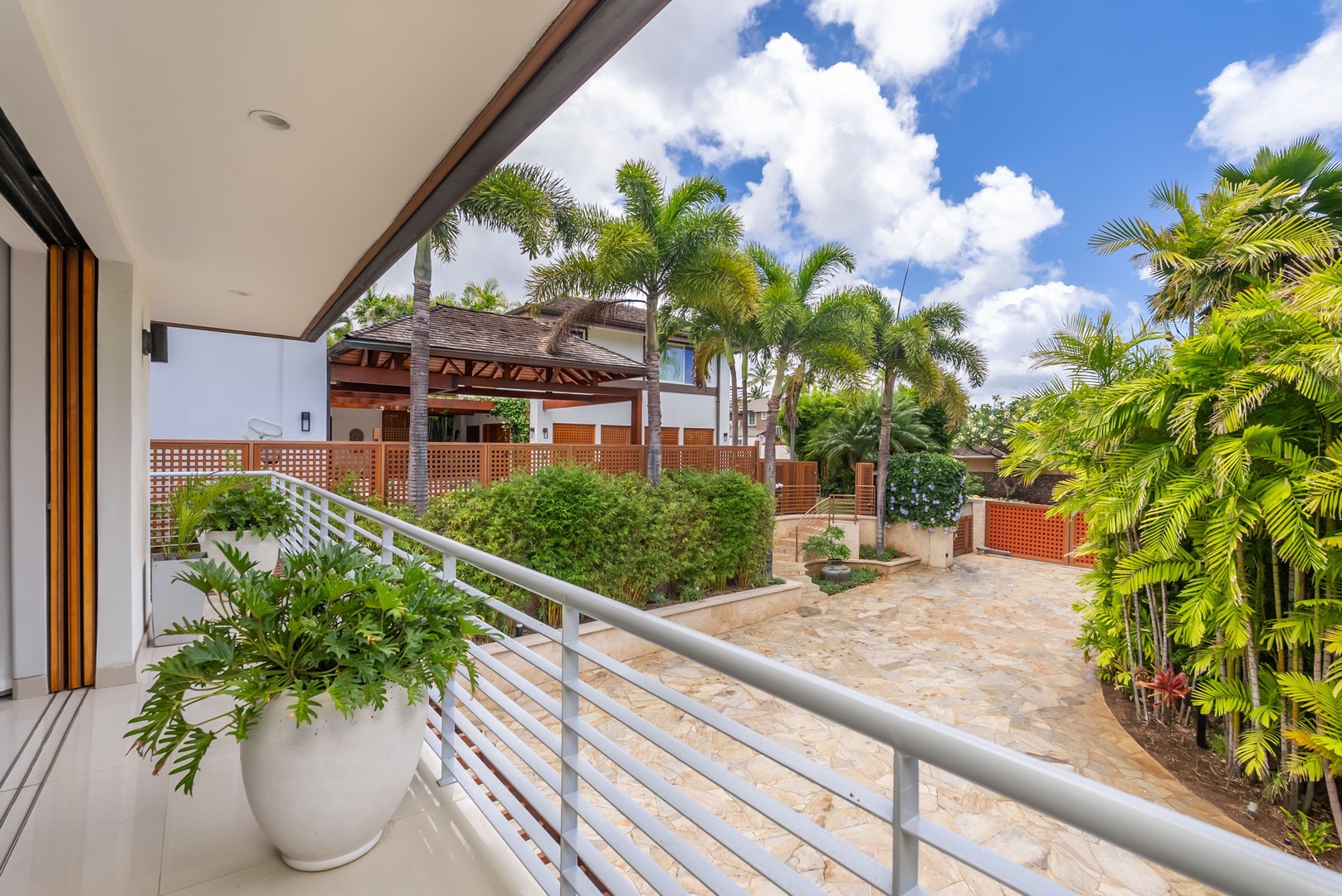 Honolulu Vacation Rentals, Diamond Head Beachfront Estate - A quiet outdoor terrace overlooks tropical palms, lush gardens, and neighboring property structures under sunny skies with scattered clouds. Perfect for relaxing outdoors during your stay.