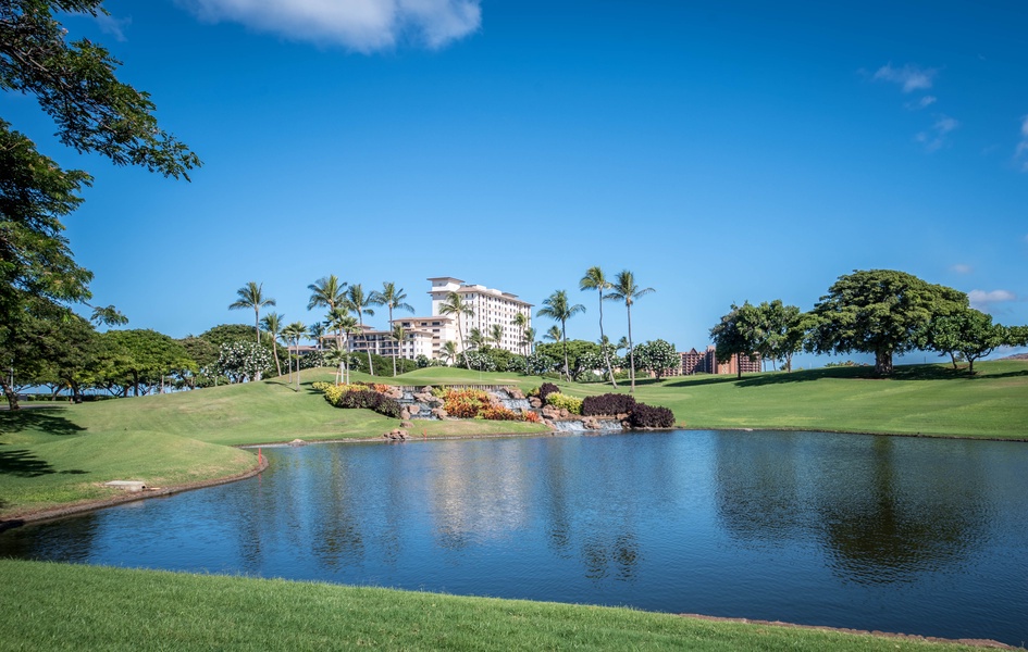 A serene resort property set amid lush landscaped grounds featuring manicured fairways, a tranquil water feature, and swaying palms under a brilliant blue sky.