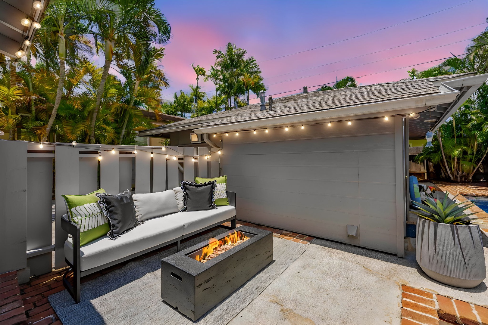 Honolulu Vacation Rentals, Ho'okipa Villa - A modern outdoor patio features a cozy lounge area with a firepit, surrounded by palm trees and illuminated by string lights beneath a colorful sunset sky.