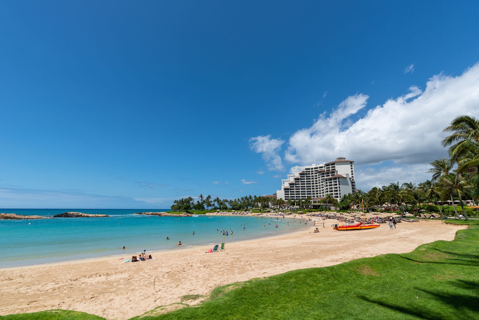 Kapolei Vacation Rentals, Ko Olina Beach Villas B604 - A pristine tropical beach stretches along turquoise waters, with palm trees and resort buildings creating an idyllic Hawaiian coastline.
