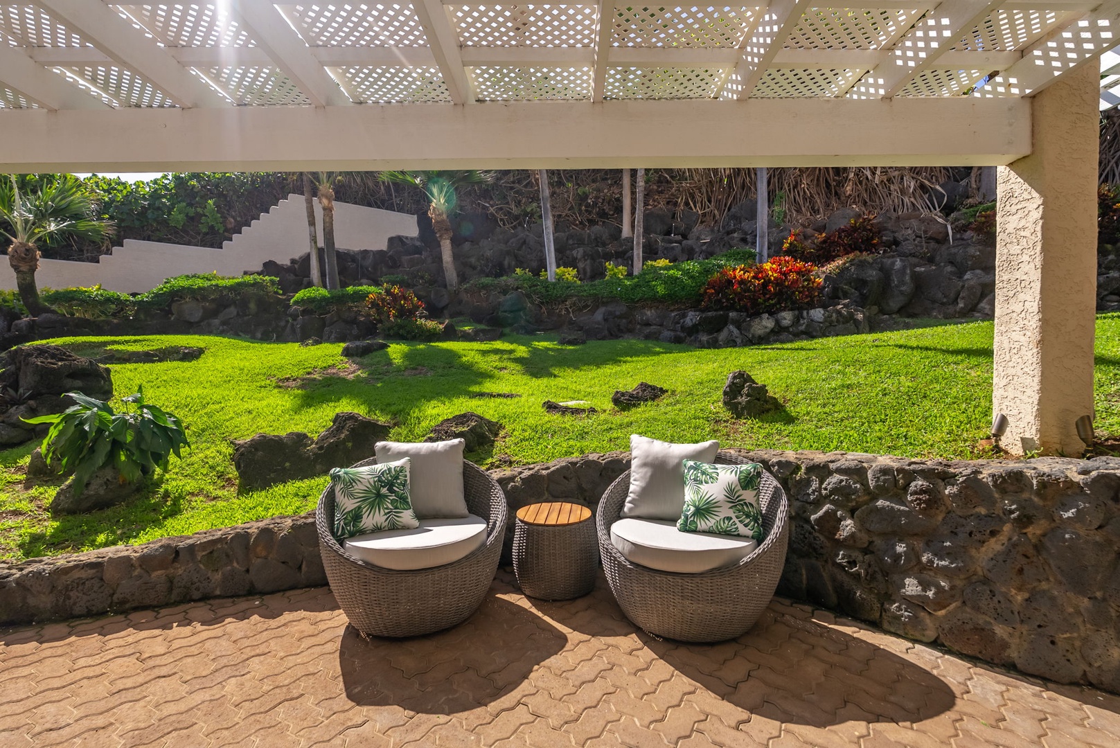 Honolulu Vacation Rentals, Black Point Ocean Villa - Garden retreat, where wicker chairs beckon beneath a pergola overlooking lush tropical landscaping and volcanic stone features.