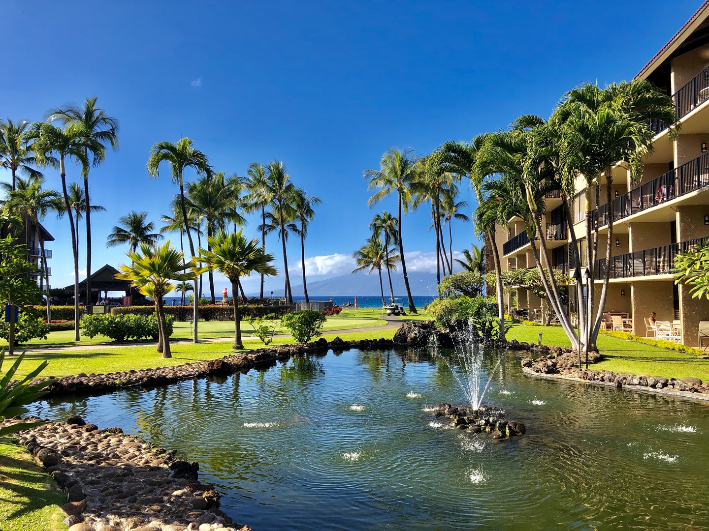 Lahaina Vacation Rentals, Papakea G-306 - A tranquil pond with a fountain enhances the tropical ambiance of the resort, offering peaceful surroundings and ocean views in the background.