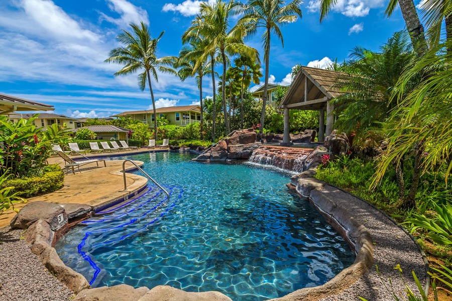 Princeville Vacation Rentals, Kaimana Villa - Tropical resort pool with waterfall feature surrounded by palm trees and lush landscaping under bright blue skies.