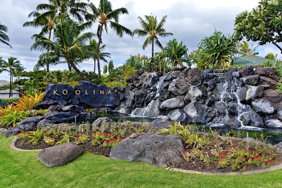 Ko Olina property entrance features a cascading waterfall over volcanic rock, surrounded by vibrant tropical gardens and swaying palm trees—a serene arrival experience in paradise.