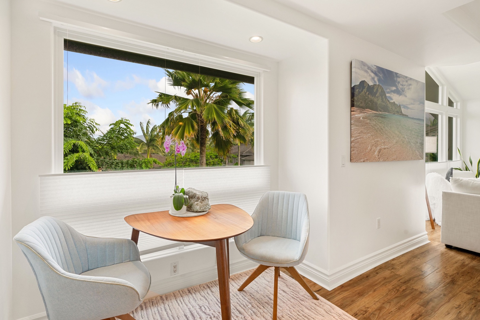 Princeville Vacation Rentals, Tropical Elegance - A serene corner of the primary suite boasts a minimalist wooden round table paired with a pastel blue upholstered chair, a perfect spot for your morning coffee w/ frames a picturesque view of lush palm trees.