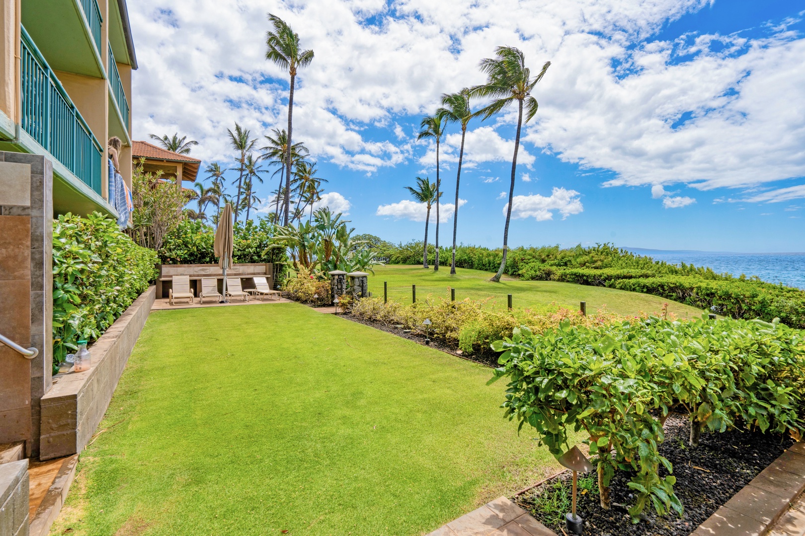 Kihei Vacation Rentals, Punahoa Beach 205 - A lounge area sits at the edge of the lawn, palms bending toward the water beyond.