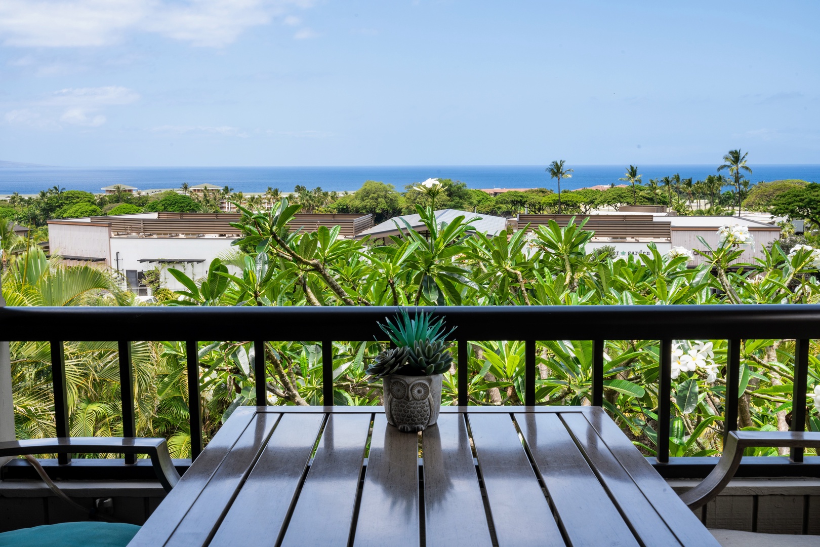 Kihei Vacation Rentals, Wailea Ekolu 204 - A stone owl planter centers the dining table, ocean visible past the plumeria and the rooftops of the shops below.