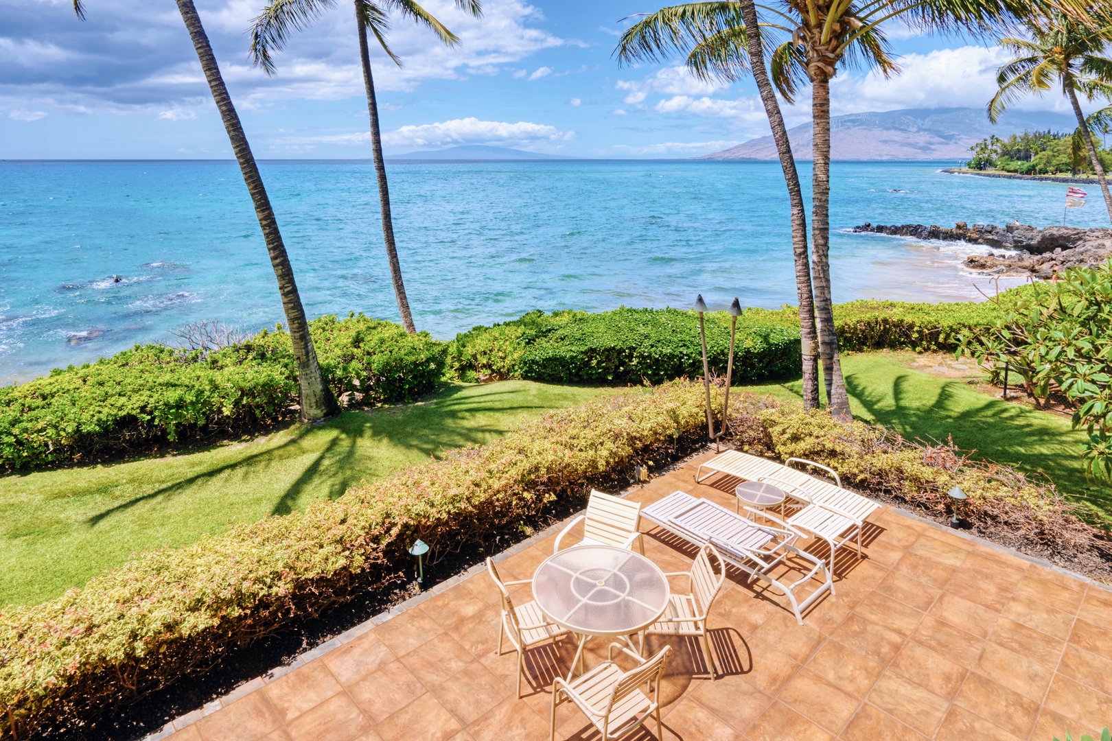 Kihei Vacation Rentals, Punahoa Beach 205 - Loungers wait beneath the palms, with a curve of beach visible just past the hedge.