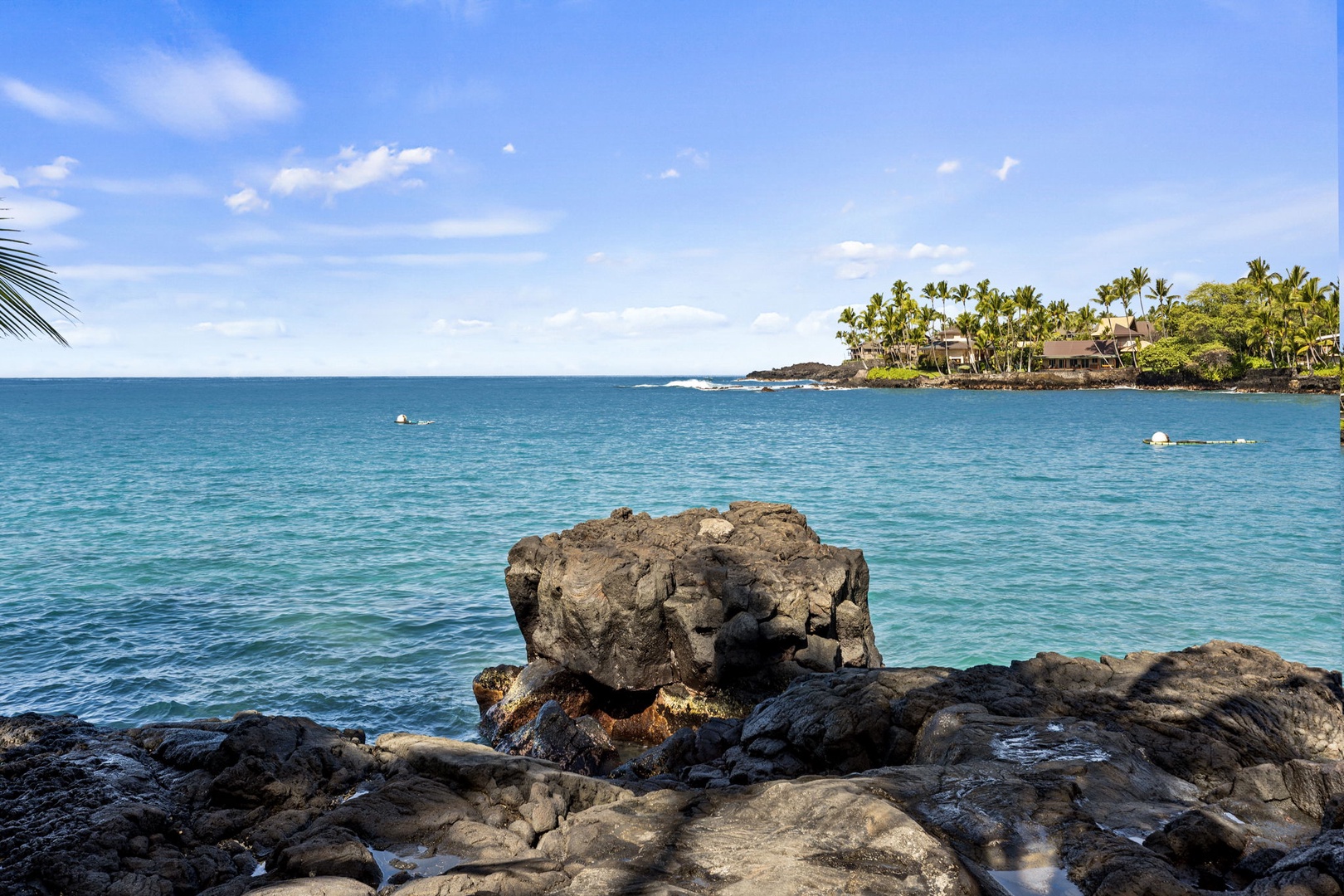 Kailua-Kona Vacation Rentals, Hahalua Bay Villa - Pristine turquoise waters meet volcanic rock coastline, with palm-fringed shores and tropical resort buildings visible across the bay.