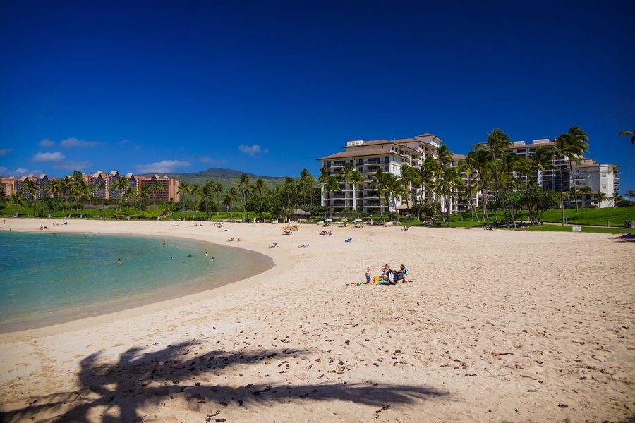A peaceful beachfront with calm turquoise waters, soft sand, and swaying palms backed by the resort buildings and lush green lawns.