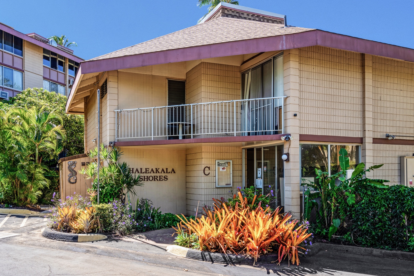 Kihei Vacation Rentals, Haleakala Shores A206 - Bright orange bromeliads and palms line the walk up to the Haleakala Shores entrance, the roofline shading the path in from the sun