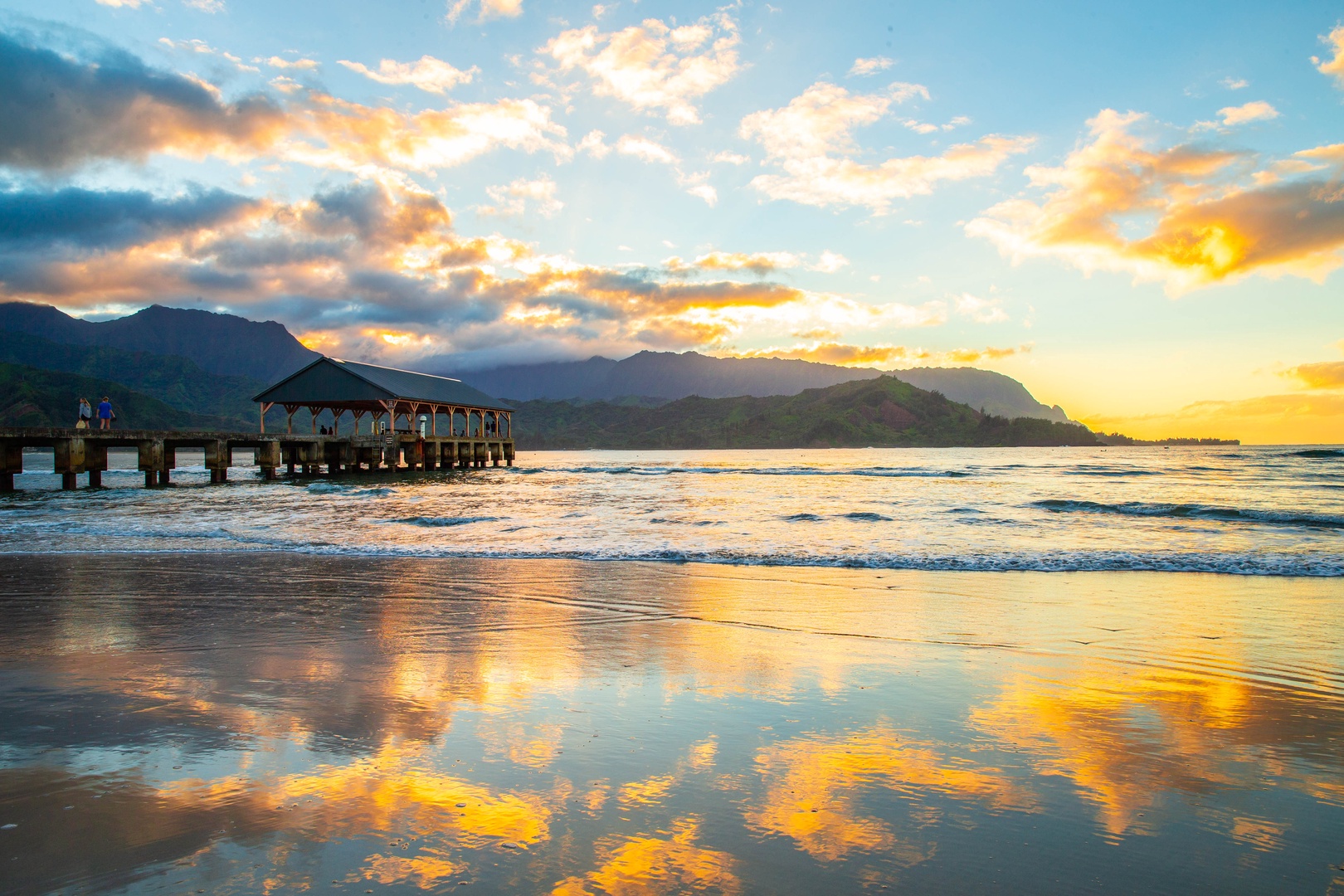 Princeville Vacation Rentals, Pohaku Kauai - Golden sunset paints the sky over pristine beach waters, with a historic pier and dramatic mountain backdrop creating an unforgettable Hawaiian paradise scene.