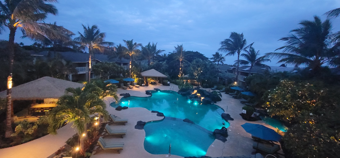 Aerial view of the pool area at twilight, surrounded by tropical palms and thatched-roof structures within lush landscaped grounds.