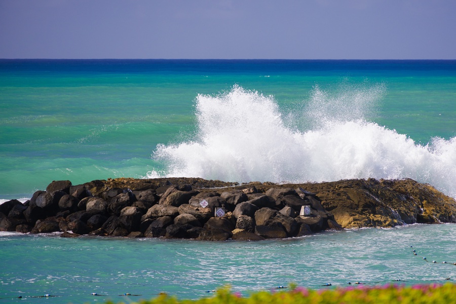 Dramatic waves crash against rocky outcroppings, with vibrant turquoise waters and clear blue skies defining the coastal landscape near the property.