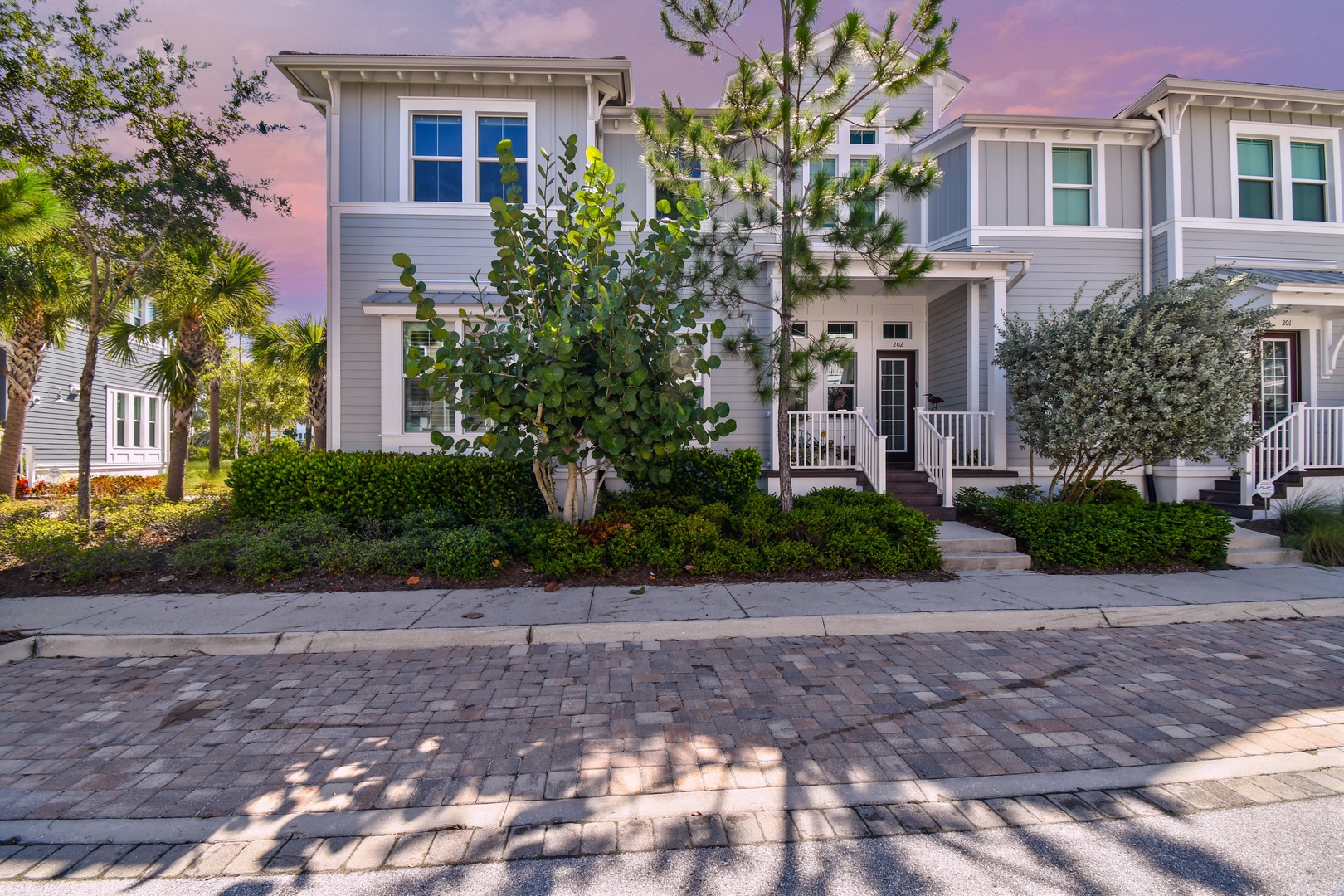 Modern coastal townhome nestled among palm trees and lush landscaping on a charming brick-paved street at dusk.