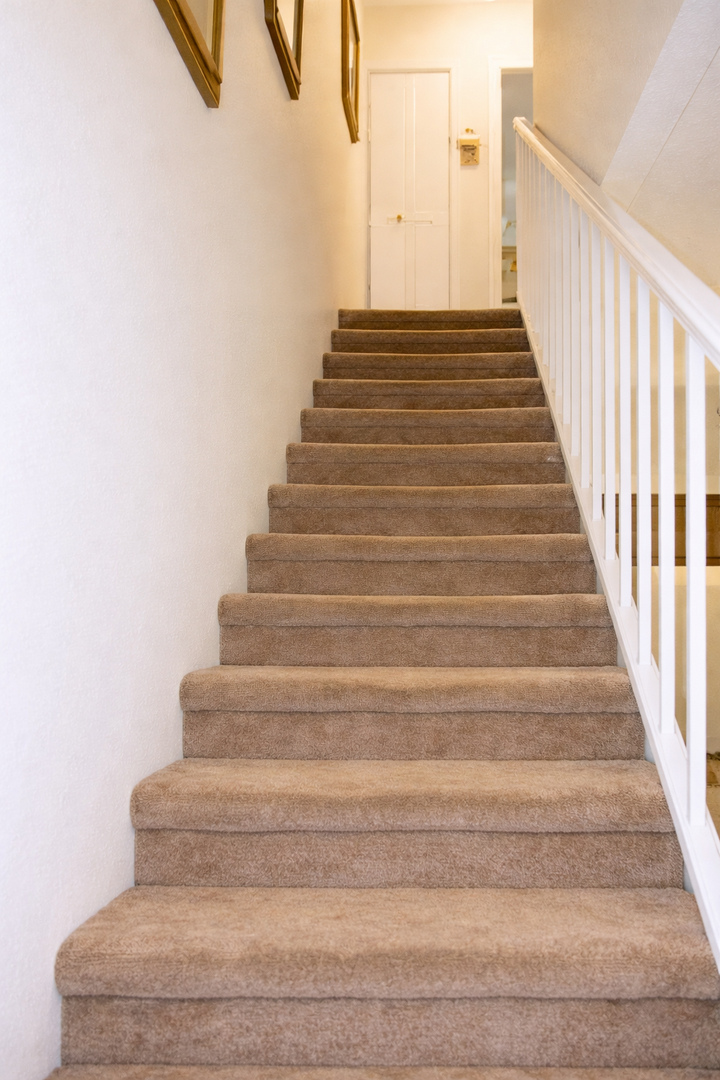 Comfortable carpeted stairs with white railings lead you to your private upstairs retreat, featuring charming exposed beams overhead.