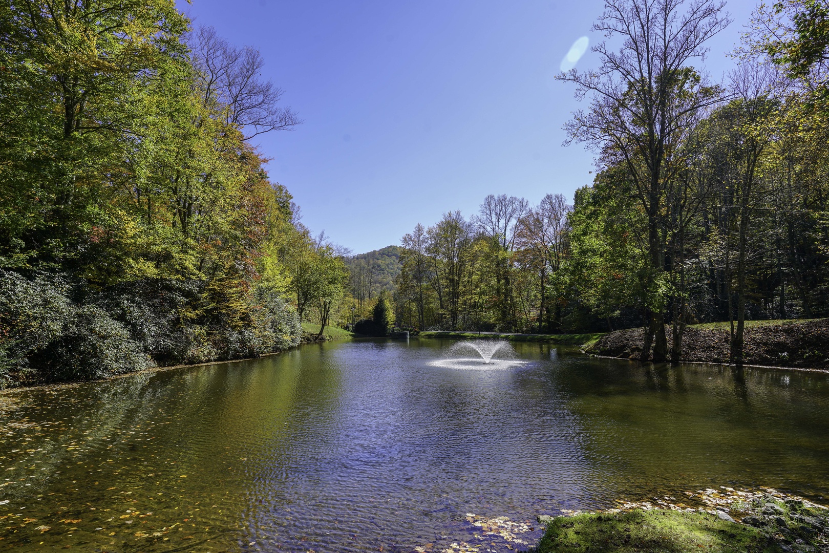 A peaceful pond nestled among lush trees and rolling hills, with a decorative fountain gracing the still water—an inviting backdrop to the property's natural surroundings.