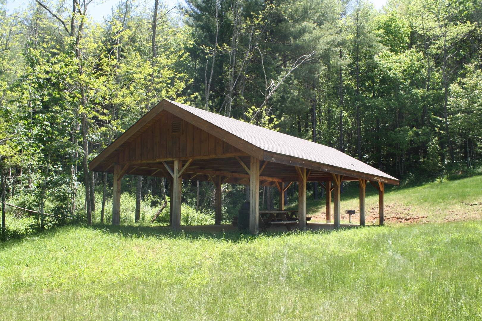 An open-air pavilion nestled in a peaceful wooded setting, offering a covered gathering space surrounded by lush greenery and gentle slopes—perfect for outdoor relaxation or group activities.