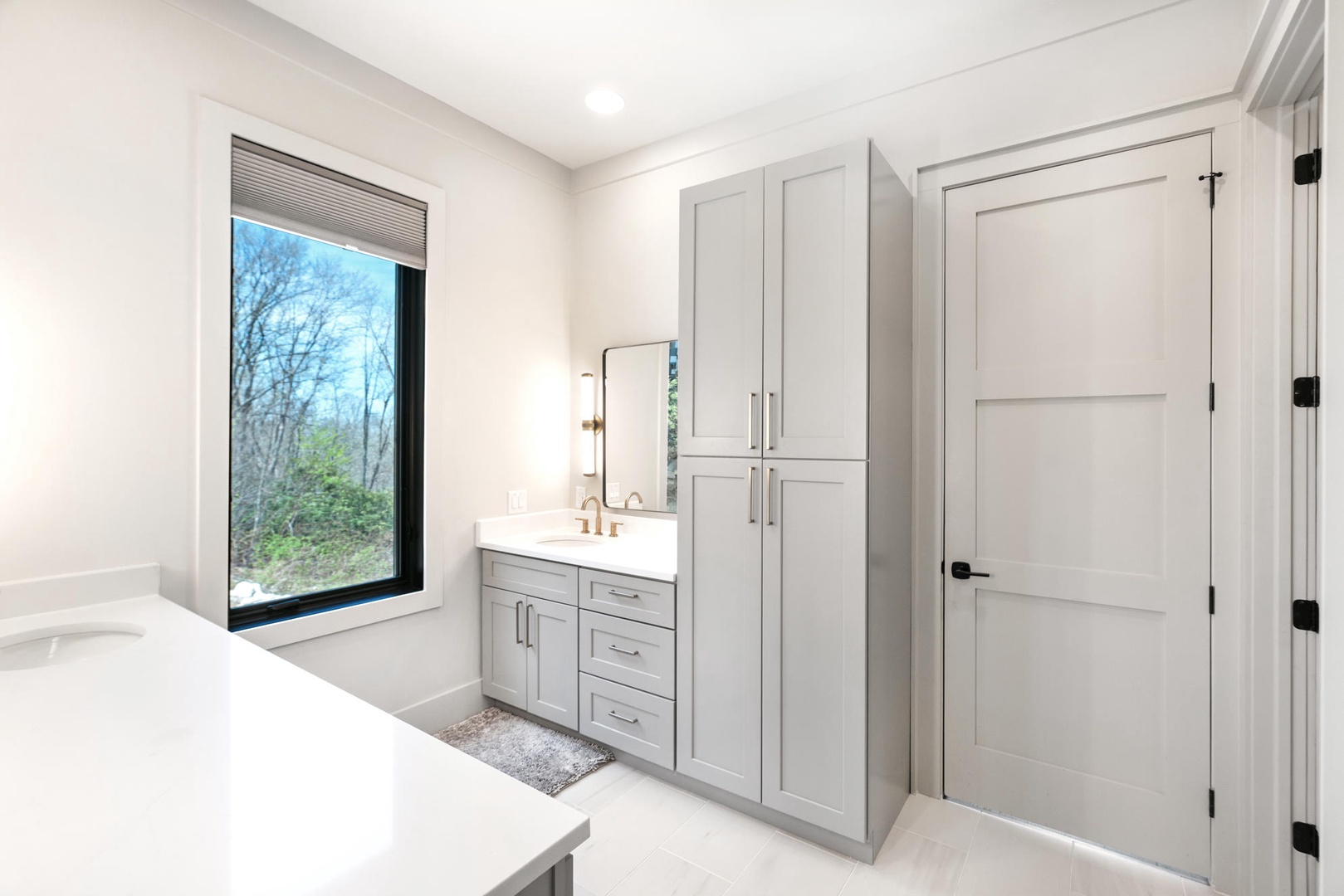 Unwind in your luxurious primary bathroom featuring elegant gray cabinetry, marble countertops, and serene forest views through the picture window.
