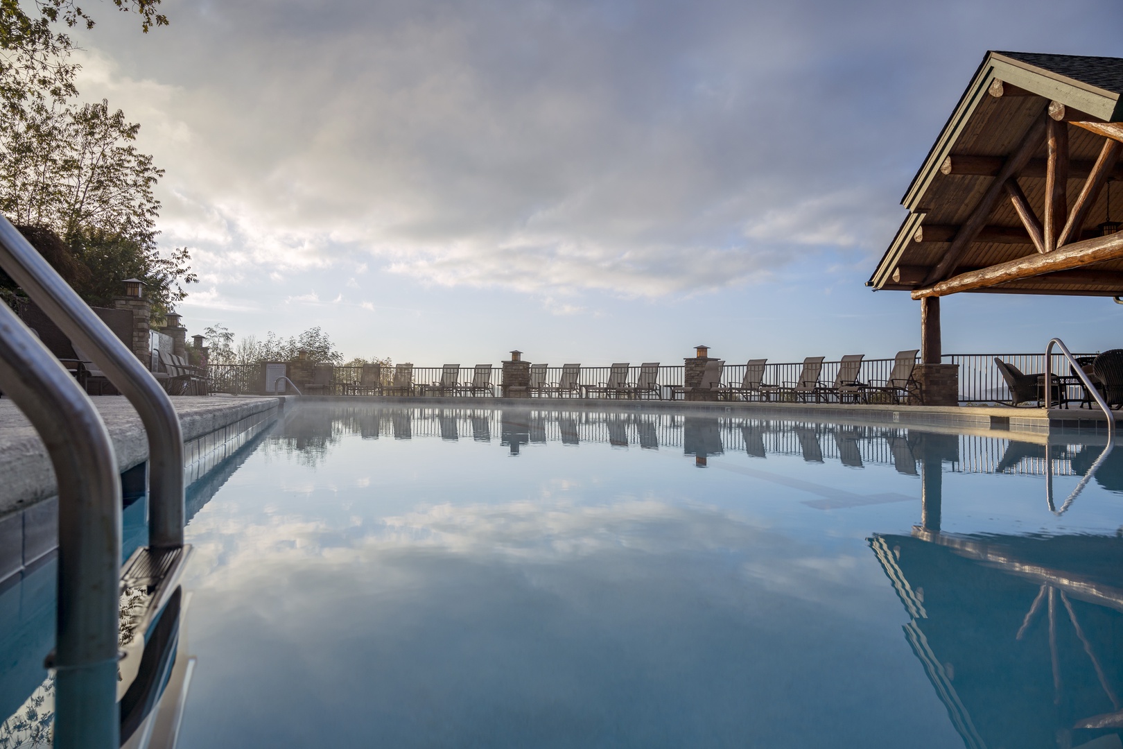 A serene outdoor pool with dramatic sky reflections, framed by a rustic timber pavilion and mountain vistas stretching beyond the deck.