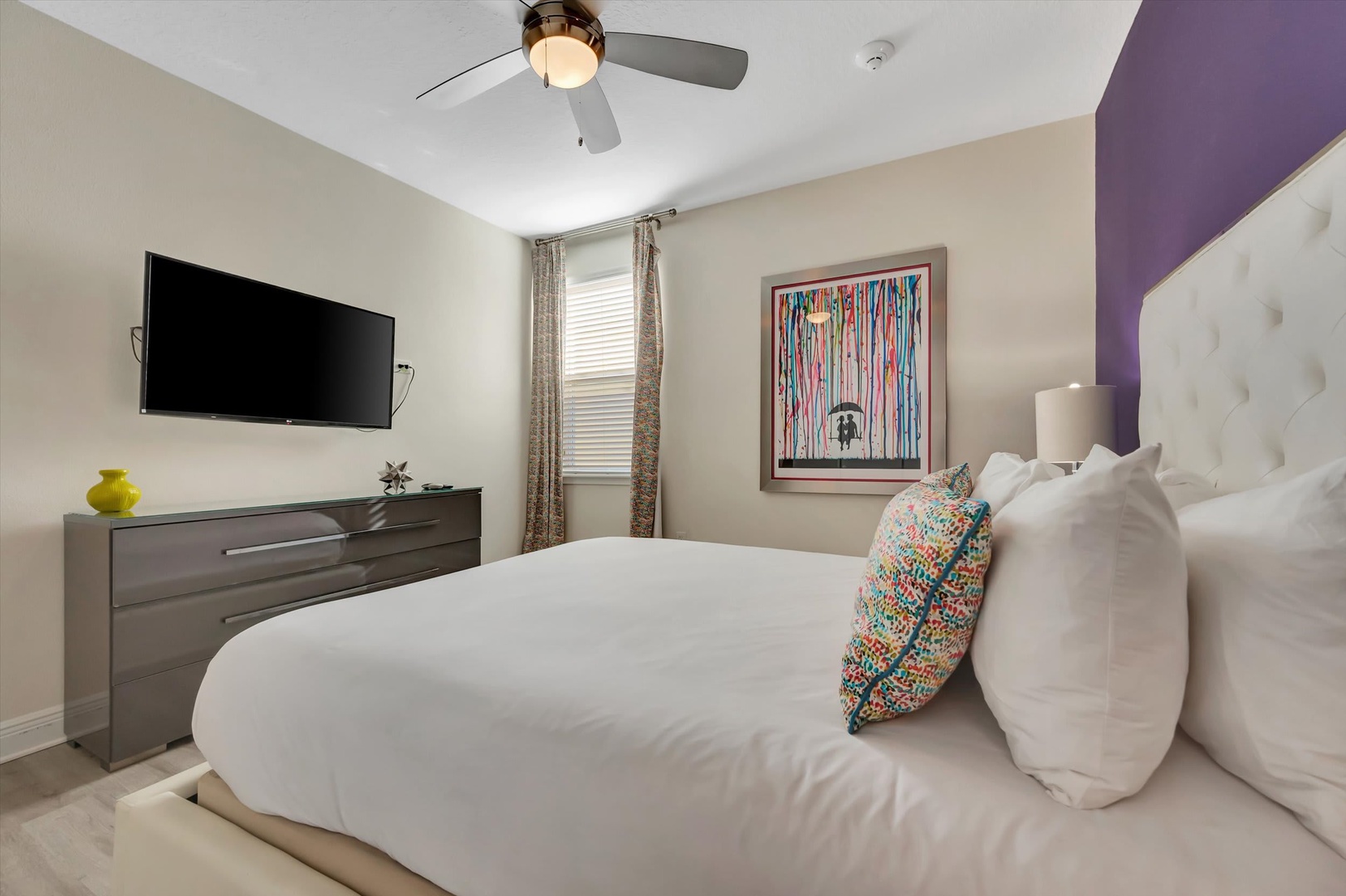 Sink into plush bedding and unwind in your stylish bedroom, complete with a ceiling fan, colorful artwork, and modern furnishings for your comfort.