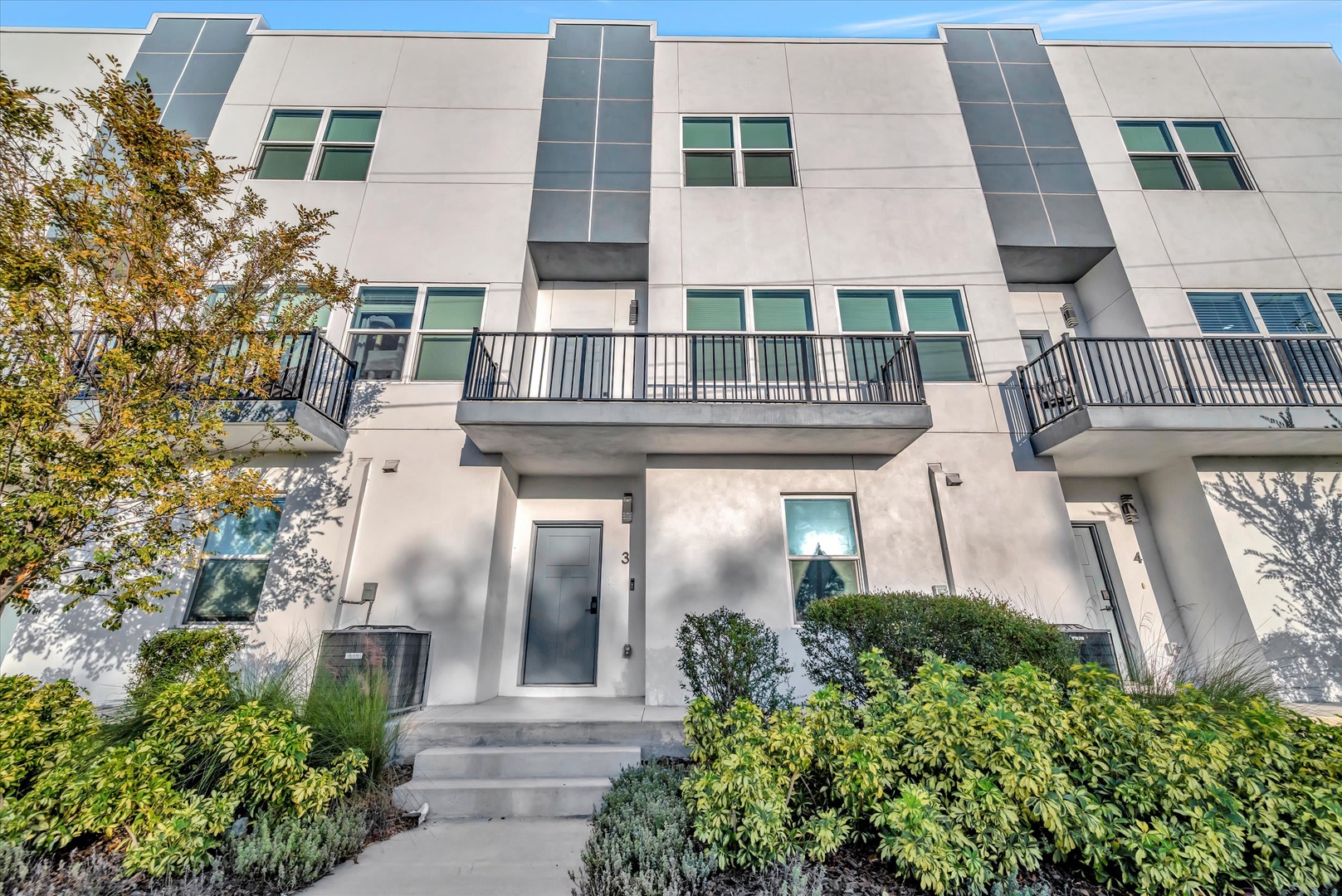 Modern townhome facade with multiple balconies, surrounded by lush landscaping and greenery, nestled in a peaceful neighborhood setting.